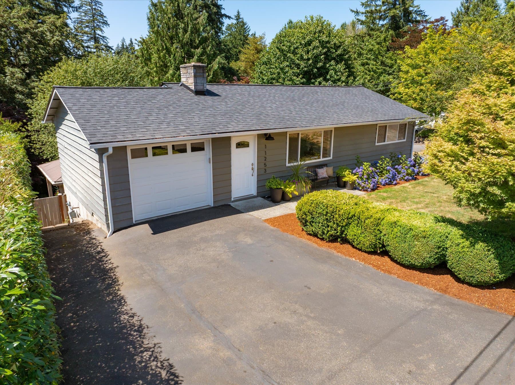 Front of the gray rambler with attached garage, driveway and clipped landscaping along the beds