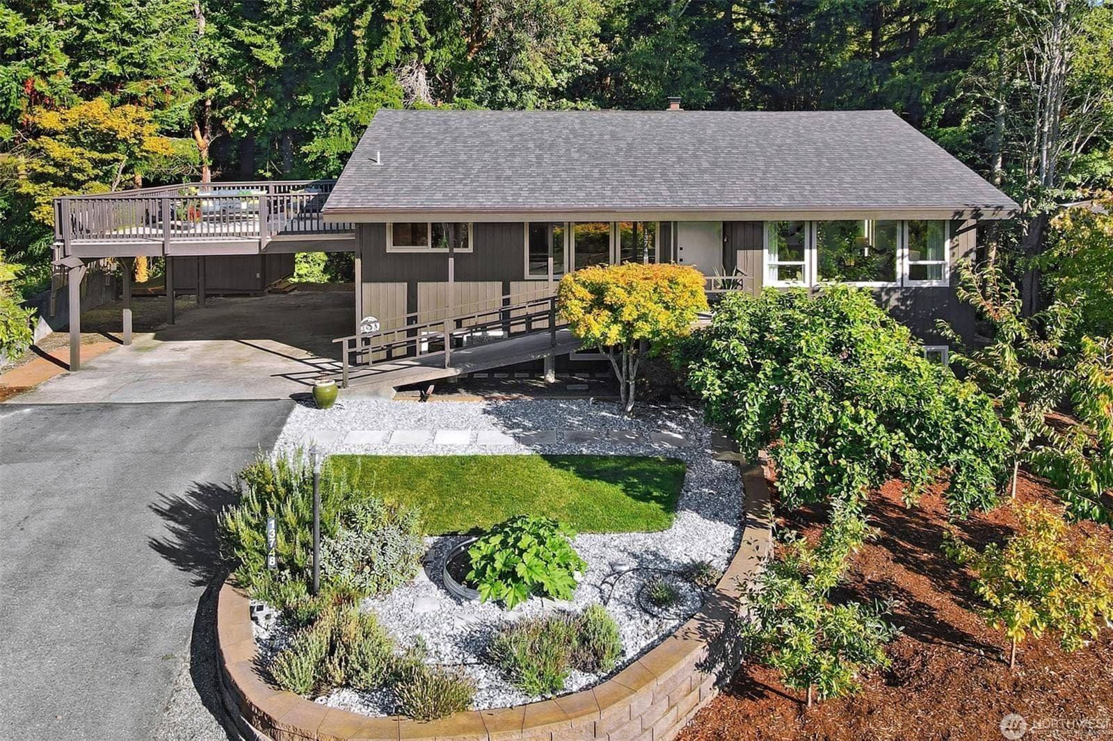 Aerial view of the brown one-story home with its wraparound deck over the carport, a landscaped front garden and tall trees behind