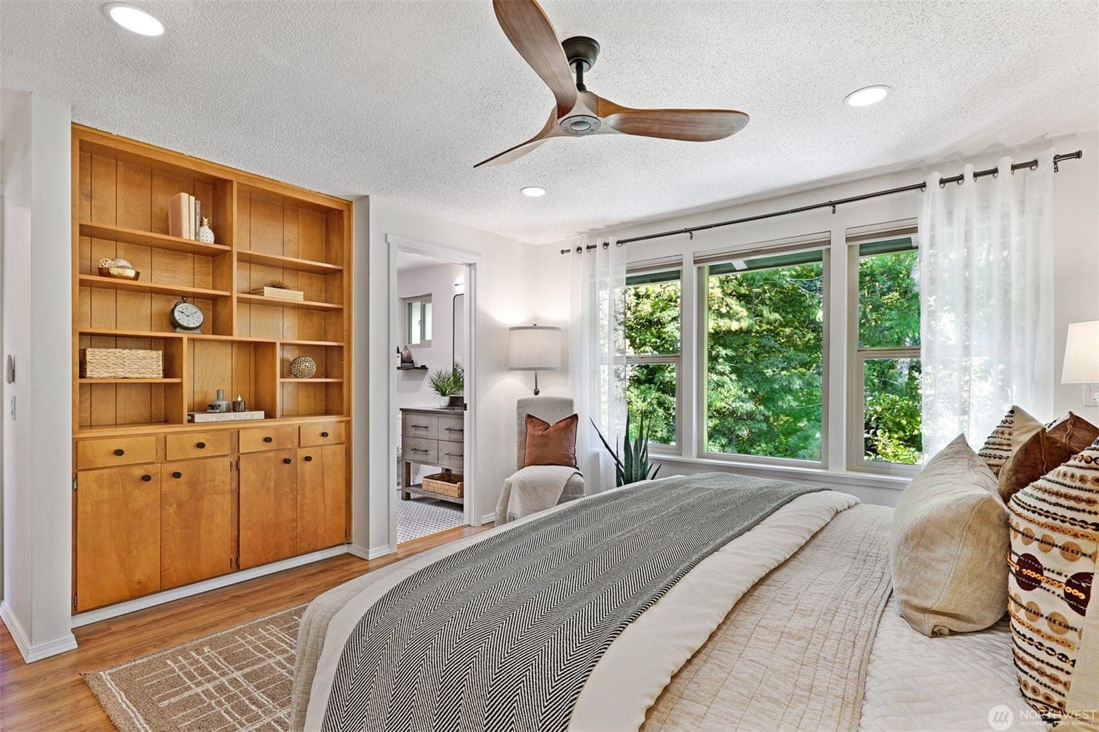 Primary bedroom with built-in wood shelving, a wall of windows onto the trees and a door through to the bath