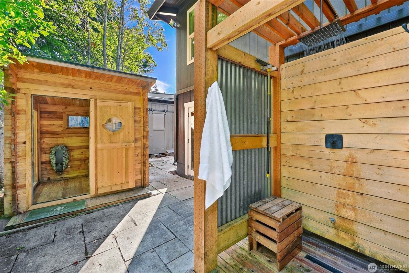 Cedar sauna with a porthole door beside an outdoor rain shower with wood and corrugated-metal walls