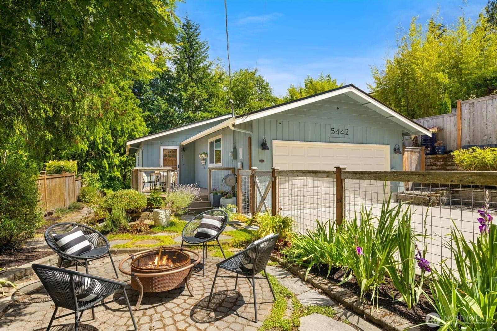 The pale-blue one-level home from the front patio, with firepit seating on pavers, planted beds and the two-car garage beyond
